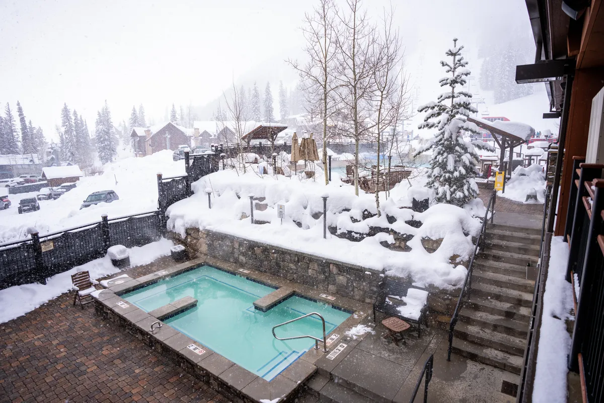A snow-covered outdoor hot tub area with steps leading down to the spa, surrounded by trees and winter scenery. People can be seen relaxing in the steaming water, while others enjoy the snow play area nearby.