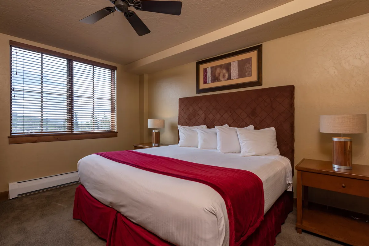 This image showcases a king-size bed adorned with plush red accents, large windows with wooden blinds that invite natural light, and tasteful artwork above the bed. The mood is tranquil and comfortable, giving a sense of a restful sanctuary.