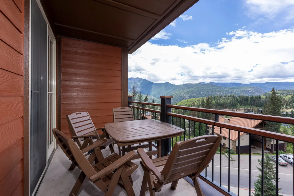 This image captures a private balcony with wooden patio furniture overlooking the dramatic San Juan Mountains. The clear sky and expansive mountain view create a tranquil setting, while small details like morning mist and the warm glow of sunrise or sunset add atmosphere. The scene invites relaxation, whether with a book, a morning coffee, or evening cocktails, with nature as the backdrop.