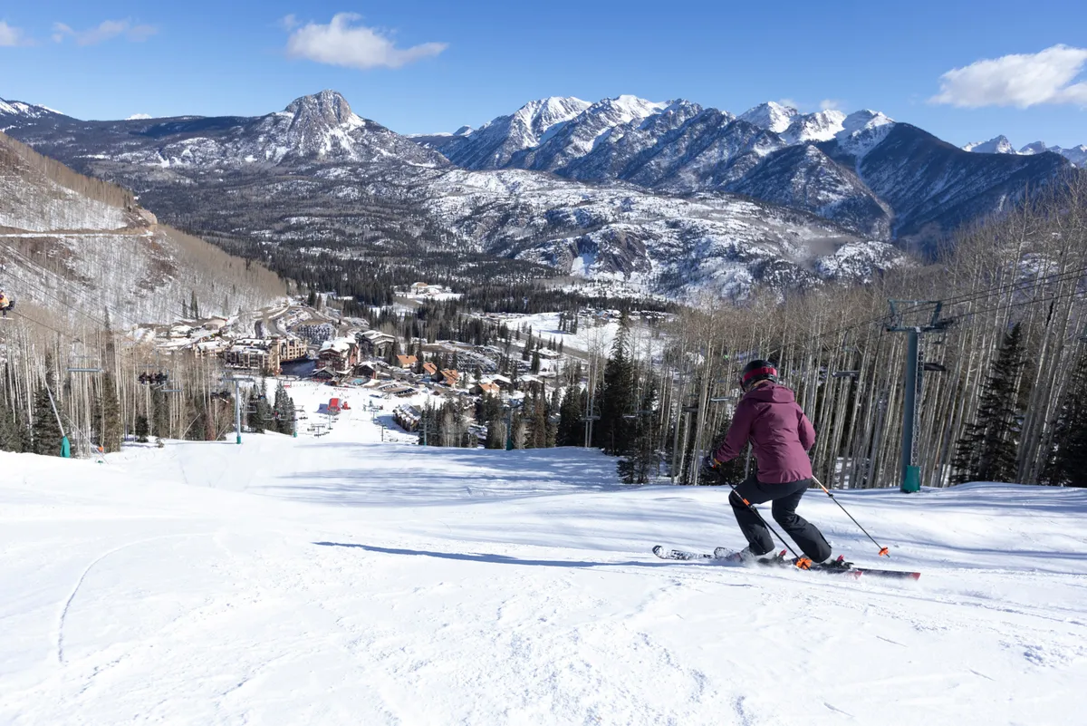 A skier in action glides down a snow-covered mountain, with the expansive ski resort and majestic peaks stretching out below. The scene is dynamic, capturing the thrill of skiing and the breathtaking winter landscape surrounding Purgatory Lodge.