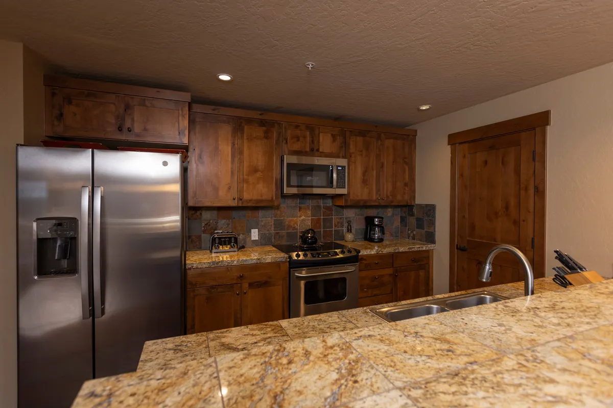 A close-up view of the kitchen's stainless steel refrigerator, granite countertop, and wooden cabinets, accompanied by appliances and hints of baking or meal prep in action. The warm ambiance suggests a space ideal for family traditions and shared experiences.
