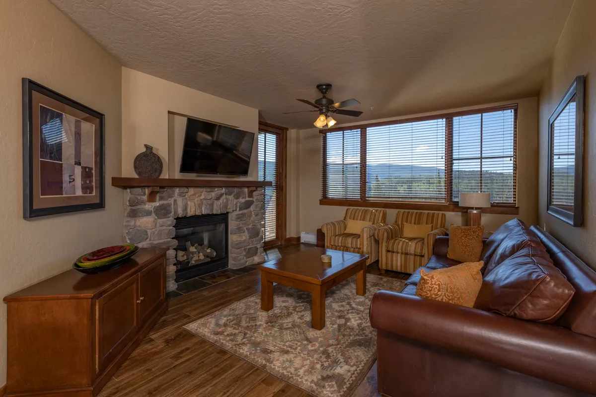 The image showcases a warm and inviting condo living room at Purgatory Lodge, featuring a stone fireplace as a central focal point. Sunlight pours through expansive windows, offering breathtaking mountain views. A plush sofa and a rustic wooden coffee table create a cozy spot for relaxing or socializing after a day on the slopes.