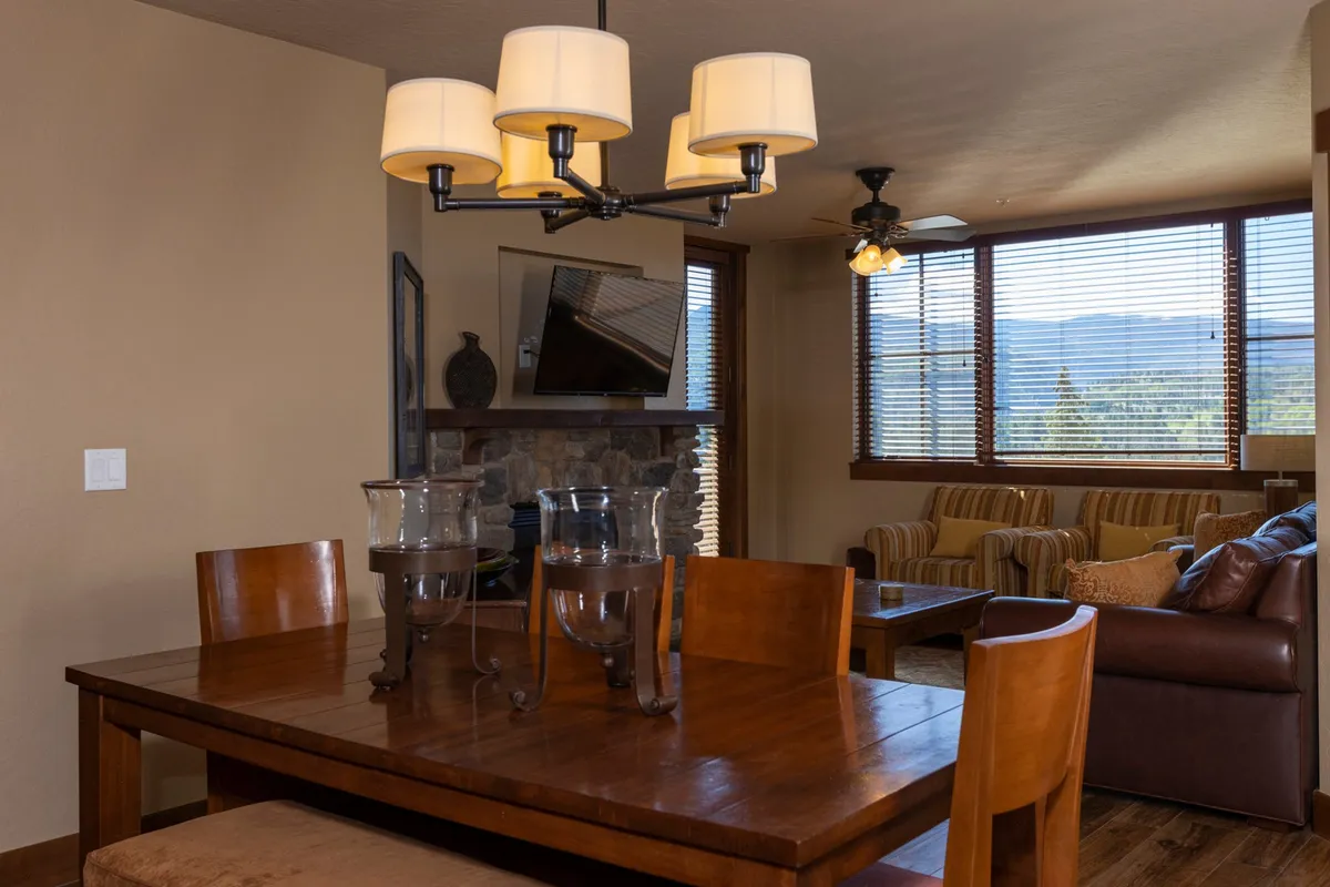 This inviting interior scene features a spacious dining table set under a glowing chandelier, with a cozy gas fireplace to the side. Large windows frame expansive mountain views, bathing the room in natural light. The setting is primed for family dinners, casual breakfasts, or gathering with friends, all anchored by plush seating and the signature lodge ambiance.